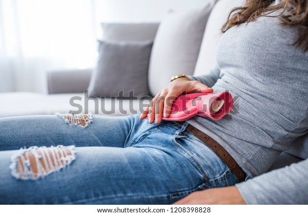 Woman in painful\
expression holding hot water bottle against belly suffering\
menstrual period pain, lying sad on home bed, having tummy cramp in\
female health concept