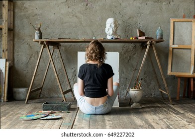 Woman Painter Sitting On The Floor In Front Of An Empty Canvas And Drawing. Art Studio Interior. Horizontal Drawing Background. Creative Concept