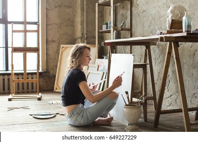 Woman painter sitting on the floor in front of the canvas and drawing. Artist studio interior. Drawing supplies, oil paints, artist brushes, canvas, frame. Workshop or art class. Creative concept - Shutterstock ID 649227229