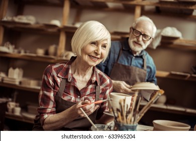 Woman Painting Clay Pot With Senior Potter At Workshop