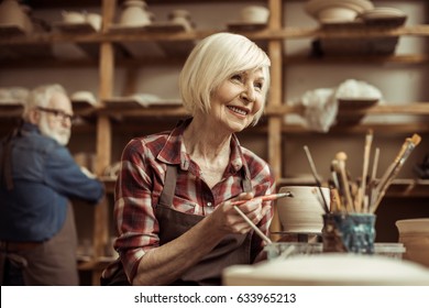 Woman Painting Clay Pot With Senior Potter At Workshop