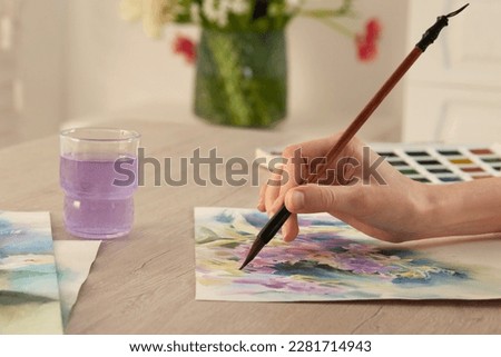 Woman painting flowers with watercolor at white wooden table indoors, closeup. Creative artwork