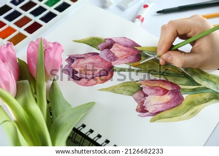 Woman painting tulips in sketchbook at white table, closeup