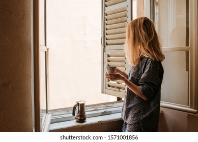 Woman in pajamas drinks morning coffee near window with shutters. Beginning of a new day. Hot drink after breakfast. Happy morning in Italy. Enjoy moment, relax lifestyle. Cozy sunnyday. Weekend vibes