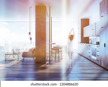 Woman In Pajamas In Stylish Kitchen With Wooden Countertops, Narrow Table And Stools. Studio Apartment Living Room On The Right. Toned Image