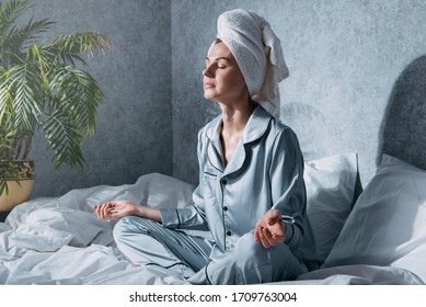 A Woman In Pajamas And A Towel On Her Head Sits On A Bed In The Morning Sun In A Lotus Position And Meditates