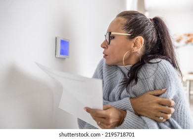 Woman With Papers In Her Hands Shivers In Sweater Looking At The Thermostat On The Wall.