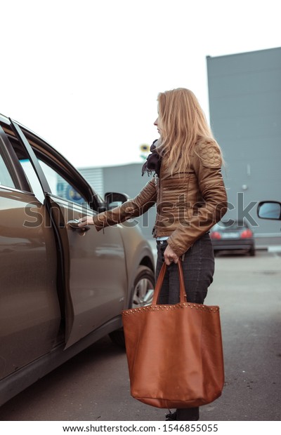 woman in parking lot of\
shopping center opens car door, in her hand is bag with groceries\
and purchases, brown leather jacket and bag, crossover car. Autumn\
in city