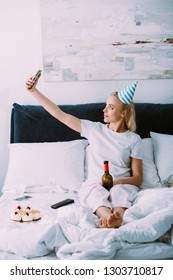 Woman In Party Hat Taking Selfie On Smartphone While Celebrating Birthday In Bed Alone