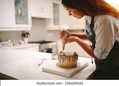 Woman Pastry Chef Decorating Chocolate Cake In The Kitchen. Female Wearing A Apron Decorating Cake With A Pastry Bag With Cream.