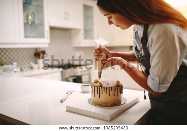 Woman pastry chef decorating chocolate cake in the\
kitchen. Female wearing a apron decorating cake with a pastry bag\
with cream.