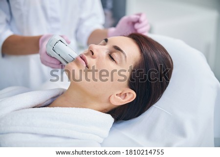 Woman patient relaxing during the ultrasonic facial treatment conducted by a cosmetician in sterile gloves