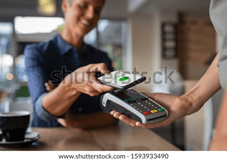 Woman paying bill through smartphone using NFC technology in a restaurant. Satisfied customer paying through mobile phone using contactless technology. Closeup hands of mobile payment at a coffee shop