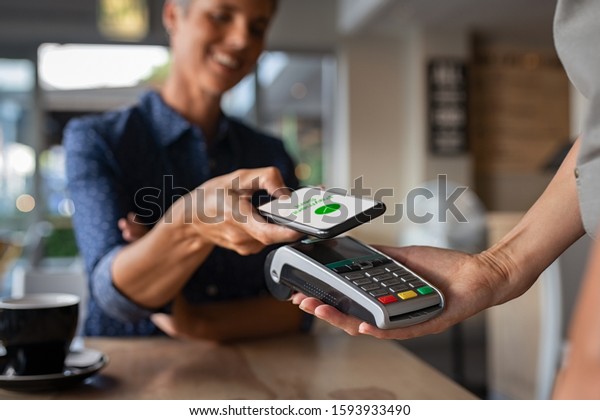 Woman paying bill through smartphone using NFC\
technology in a restaurant. Satisfied customer paying through\
mobile phone using contactless technology. Closeup hands of mobile\
payment at a coffee shop