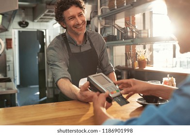 Woman Paying By Credit Card And Entering Pin Code On Reader Holded By Smiling Barista In Cafeteria. Customer Using Credit Card For Payment. Mature Cashier Accepting Payment Over Nfc Technology.