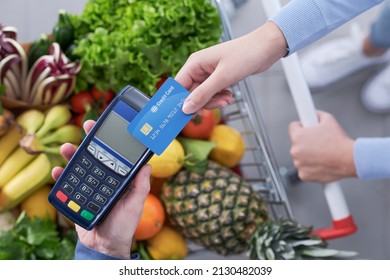 Woman Paying For Groceries Using Her Credit Card, The Supermarket Cashier Is Holding The POS Terminal