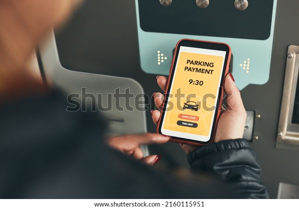 Woman paying for parking ticket at car parking\
payment machine using mobile app on smartphone. Driver using\
smartphone to pay for parking. Car park application on mobile\
phone. Mobile payment app