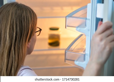Woman peers into a refrigerator and only sees a jar of pickles in fridge. Woman inspecting almost empty fridge at home during self-isolation. Concept of diet and quarantine at covid-19 coronavirus 
