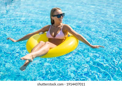 Woman With Perfect Sexy Body In Stylish Black Bikini And Glamorous Sunglasses, Posing On An Inflatable Yellow Ring In The Swimming Pool Outdoors