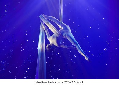 A woman performing aerial silks in a split position with her legs stretched out upside down, illuminated by blue light - Powered by Shutterstock