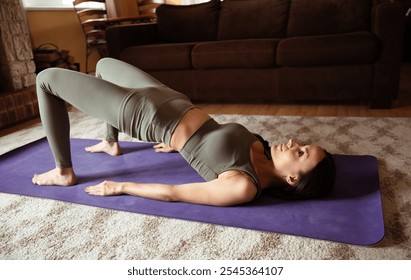 A woman performing a bridge pose on a purple yoga mat, wearing green activewear, indoors in a cozy living room with warm lighting, a sofa, and a peaceful atmosphere. - Powered by Shutterstock