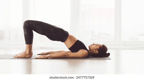 A woman is performing a yoga bridge pose on a mat in a well-lit studio with large windows. She is focused and appears to be in the middle of a workout session. - Powered by Shutterstock