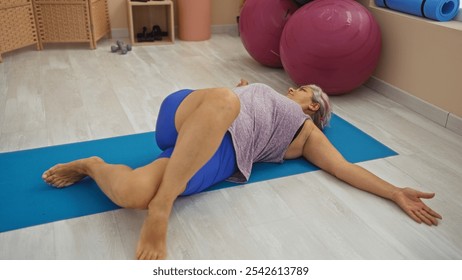 Woman performing a yoga stretch indoors on a blue mat in a well-lit gym surrounded by exercise balls and equipment. - Powered by Shutterstock