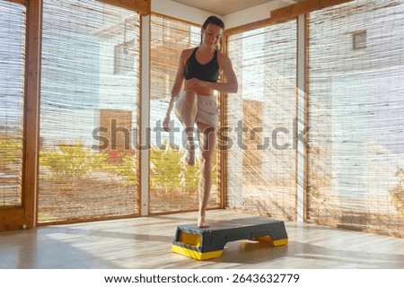 A woman performs an exercise routine on a step platform in a bright indoor space, surrounded by scenic outdoor views.