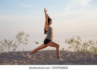 A woman performs a yoga lunge pose on a serene sandy beach during a calm morning, showcasing balance, flexibility, and mindfulness under soft sunlight with scenic natural surroundings - Powered by Shutterstock