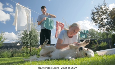 Woman petting a Golden Retriever on the grass while a man hangs clothes on a line in a sunny garden. High quality photo - Powered by Shutterstock