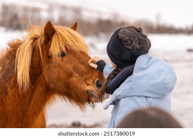 A woman is petting a horse with a brown mane. The woman is wearing a blue jacket and a black hat - Powered by Shutterstock