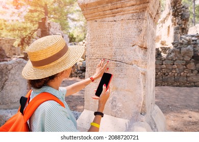 Woman philologist and tourist with smartphone reads and translates ancient Greek from columns in the ruins of an antique city. Linguistics and archaeology