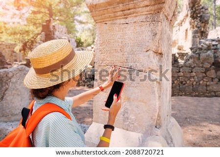 Woman philologist and tourist with smartphone reads and translates ancient Greek from columns in the ruins of an antique city. Linguistics and archaeology