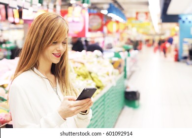 Woman With Phone In Grocery Aisle. Hipster Girl Using Mobile In Store Aisle. Shopping App Concept.