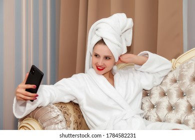 Woman With Phone On Sofa Of Hotel Room After Shower. Pretty Female Of Slavic Appearance In Dressing Gown And Towel On Her Head. Happy Lady. Lesson With Smartphone. Cute Girl After Washed