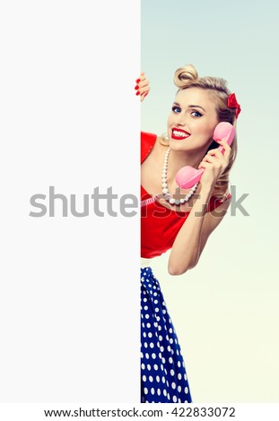 woman with phone, in pin-up style dress, showing blank signboard with copyspace area. Caucasian blond model posing in retro fashion and vintage concept studio shoot.