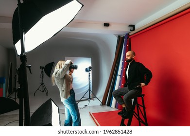 Woman Photographer Making Business Portraits For Handsome Bearded Man On Red Background In Photo Studio. Work Of A Photographer. Backstage Photo