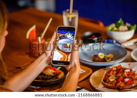 Woman photographing dinner in restaurant through mobile phone at table. Hand hold smartphone taking photo of dish before eating in cafe. Phone photo of food for social media or blogging