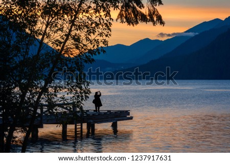 Woman Photographing a Dramatic Sunset on Lake Crescent, Washington. Using an cell phone a woman captures a beautiful sunset over the Olympic Mountains while standing on a pier at lake Crescent Lodge.