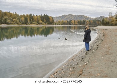 Woman photographing ducks along a tranquil lake shore with autumn forest reflections in scenic landscape. - Powered by Shutterstock