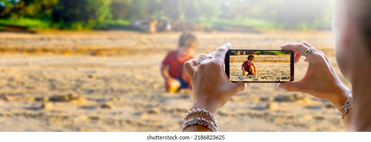 Woman photographing her little son with a cell phone. The child is playing in the sand on the beach. Camera of a smartphone. - Powered by Shutterstock