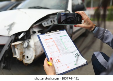 Woman photographs a broken car on smartphone and holds insurance documents in her hands. Damage assessment after car accident concept
