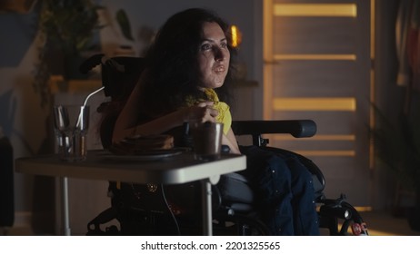 Woman With Physical Disability Sitting In Motorized Wheelchair In Front Of TV And Relaxing At Home At Night Watching TV Show