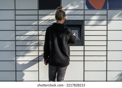 Woman Picking Up A Package From A Smart Electronic Steel Parcel Locker Box, Automatic Mailboxes. Paczkomat Delivery Service, Collection Machine. Using App On Her Phone To Open The Door.