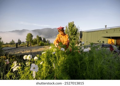 A woman picks flowers in the early morning light, surrounded by a misty mountain landscape, with a modern cabin and an electric car in the background - Powered by Shutterstock