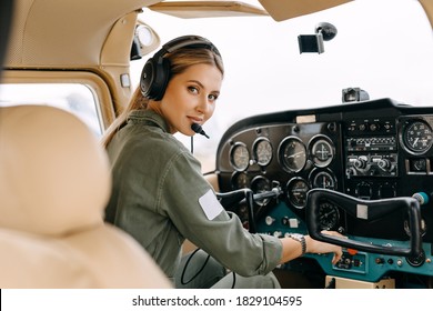 Woman pilot sitting in private airplane cockpit, wearing headset, looking at camera, smiling.