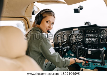 Woman pilot sitting in private airplane cockpit, wearing headset, looking at camera, smiling.