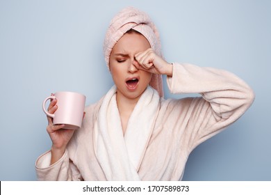 Woman In Pink Bathrobe With Towel On Head Holding Cup Of Tea Or Coffee In Hand, After Shower At Morning, Sleepy And Tired, Wants To Sleep, Rubbing Her Eyes And Yawning. Shift Work Sleep Disorder