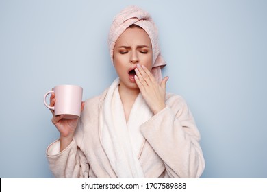 Woman In Pink Bathrobe With Towel On Head Holding Cup Of Tea Or Coffee In Hand, After Shower At Morning, Sleepy And Tired, Wants To Sleep, Rubbing Her Eyes And Yawning. Night Owl Type Concept