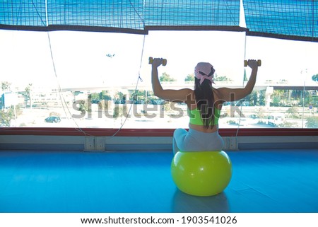 Woman with pink cancer scarf lifting weights sitting on yellow pilates ball in a gym, in front of a large window and with blue parquet floor.